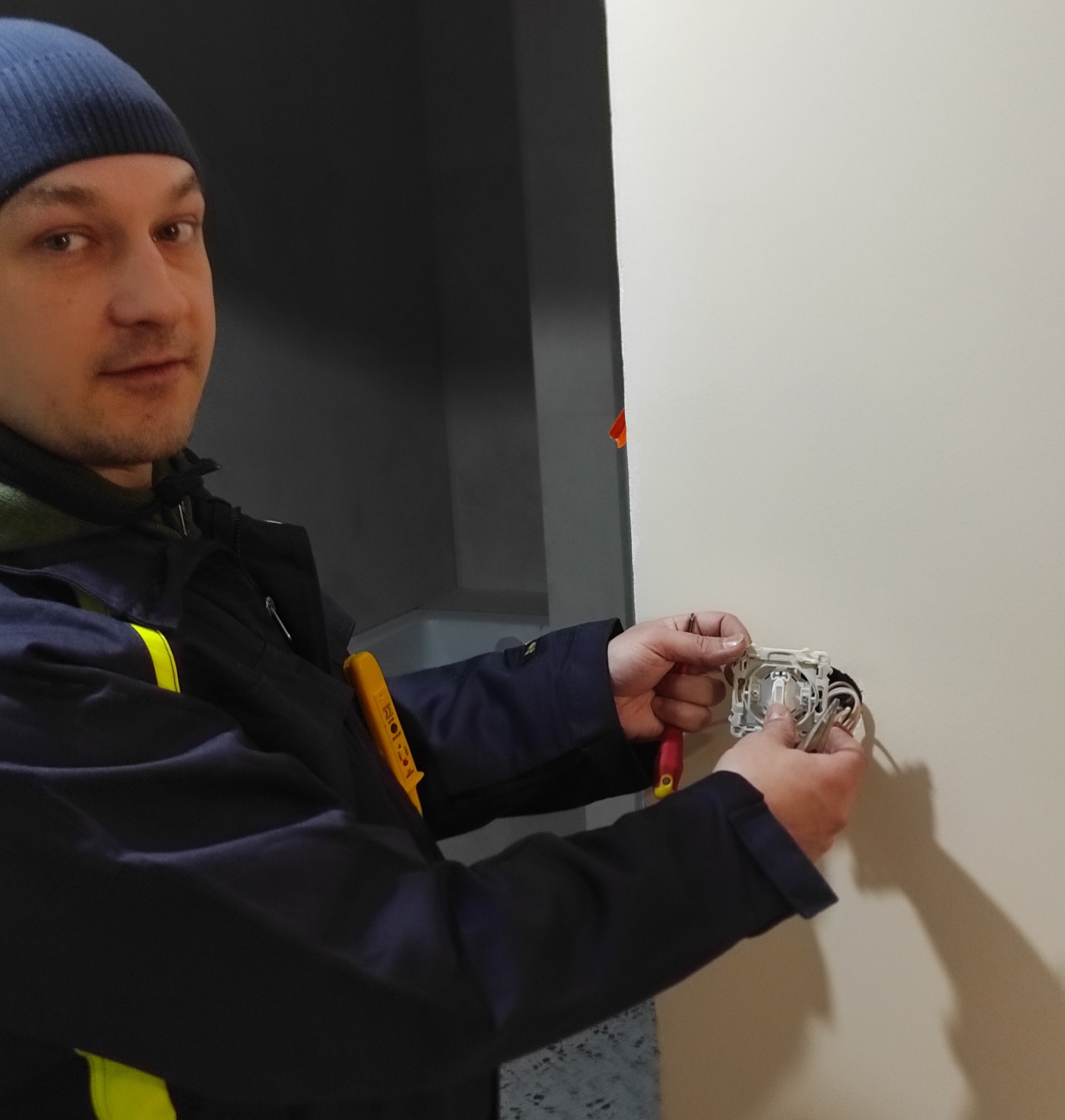 Elektryk w niebieskiej czapce i kombinezonie montuje gniazdko elektryczne, używając śrubokręta. W tle widoczna ściana i fragment pomieszczenia.