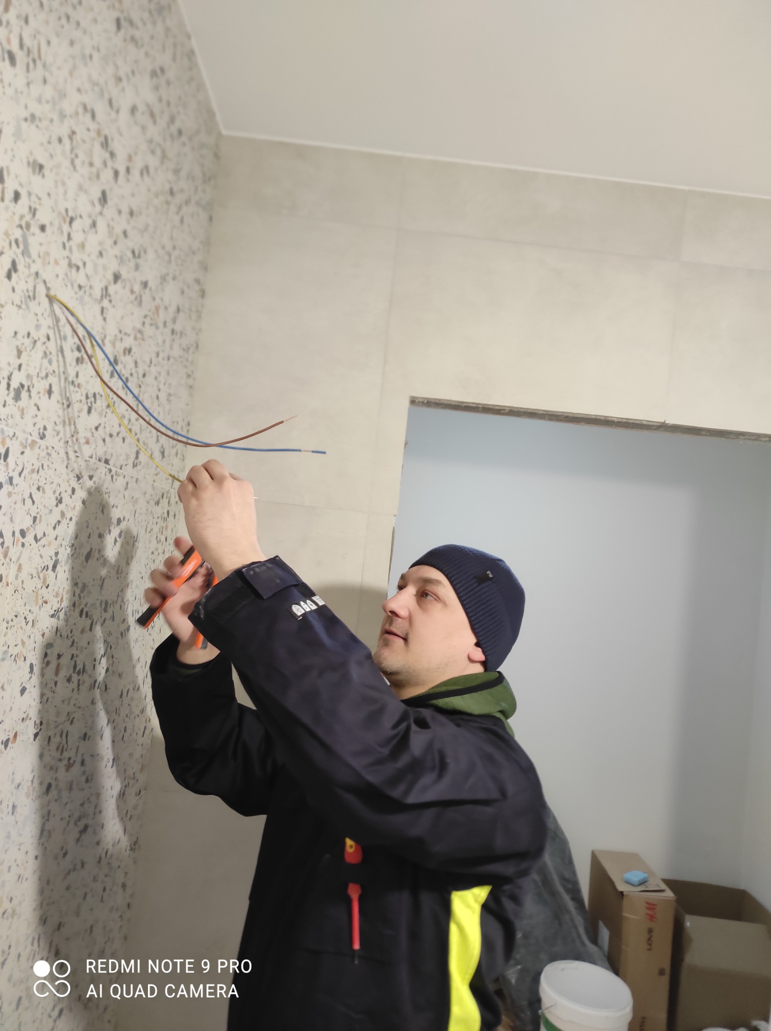 Elektryk w czapce, w trakcie pracy z przewodami na ścianie wykończonej mozaiką, używa kombinerek. Widoczne kable w kolorach: niebieski, żółty i brązowy.