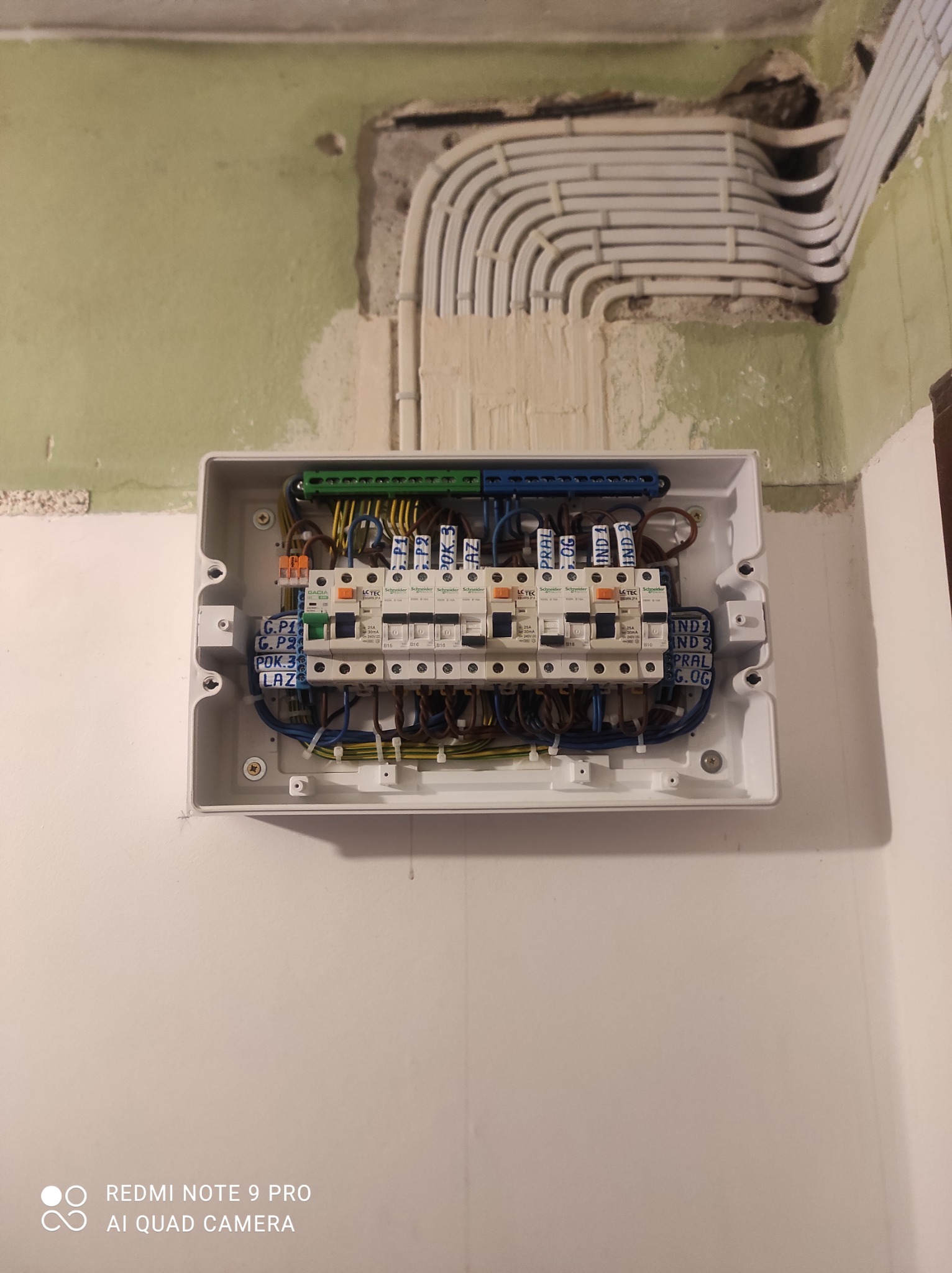 Otwarta skrzynka bezpiecznikowa z zainstalowanymi bezpiecznikami i okablowaniem na ścianie. Nad skrzynką widoczne rury instalacyjne w ścianie.