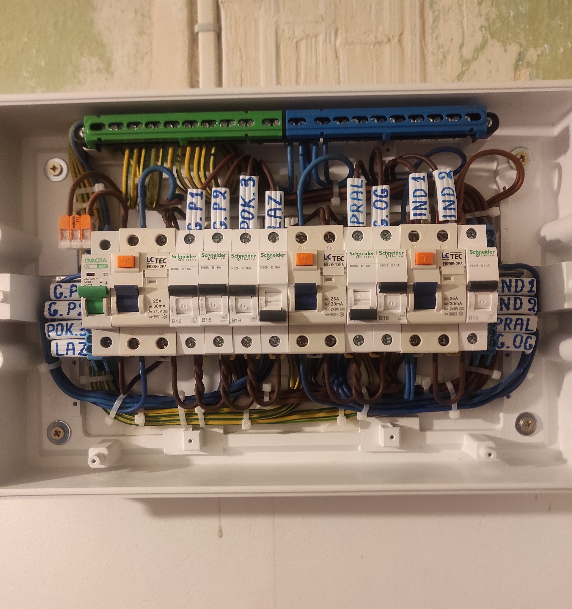 Rozdzielnica elektryczna z bezpiecznikami, okablowaniem i listwami zaciskowymi w białej obudowie. Widoczne opisy przewodów i oznaczenia zabezpieczeń.