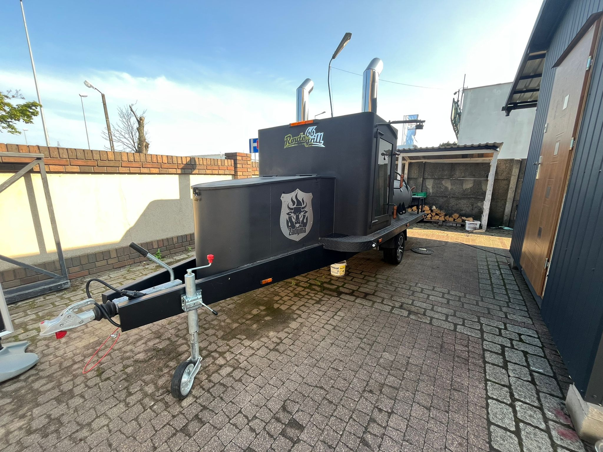 Mobilny smoker Route 66 Grill z logiem Zadymia, na kostce brukowej, z drewnem w tle. Urządzenie do wędzenia i pieczenia mięs, gotowe do użycia na imprezie plenerowej.