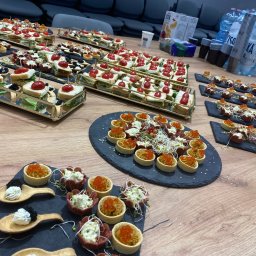 FUNDACJA PANA JAJKO - Elegancko podane przekąski na drewnianym stole: mini kanapeczki z pomidorkami, tartaletki z kawiorem i pastami, ułożone na złotych tacach i czarnych łupkach.