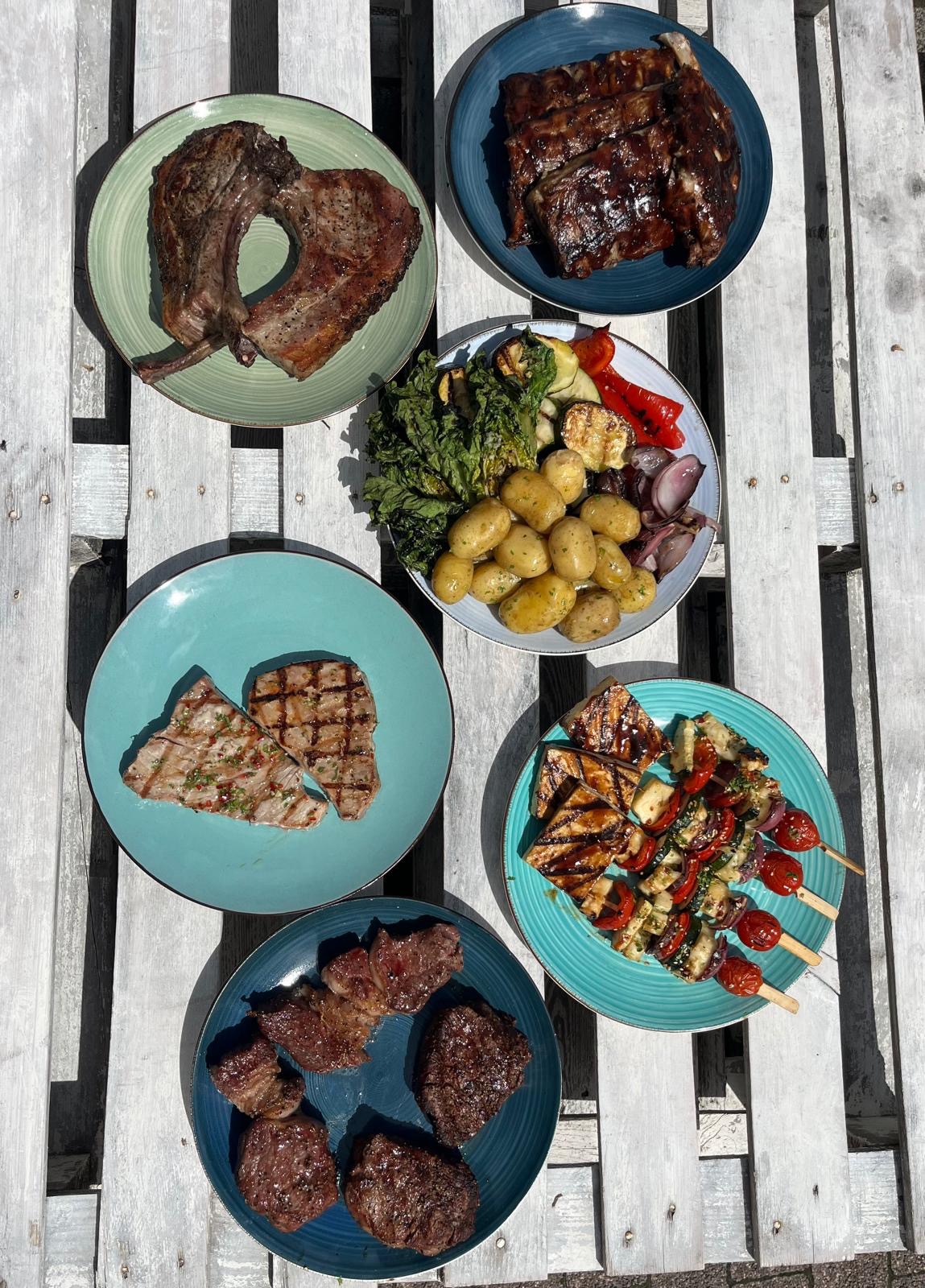Zestaw talerzy z różnorodnymi daniami grillowanymi: mięsa, warzywa, szaszłyki, ziemniaki. Kompozycja na drewnianym stole. Catering z Gostynia.