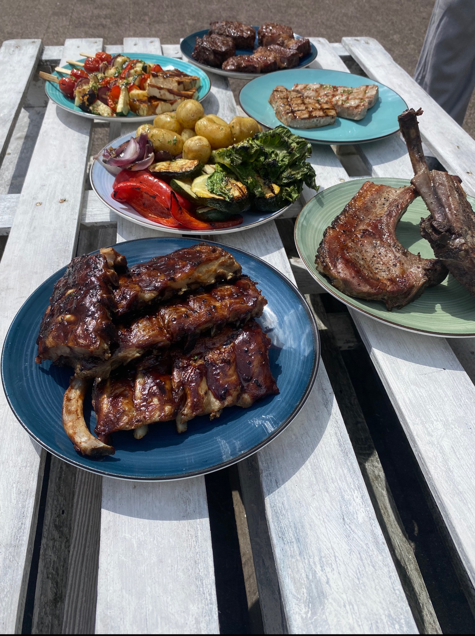 Grillowane żeberka BBQ, szaszłyki warzywne i mięsa na talerzach, ułożone na białym, drewnianym stole w plenerze. Różnorodność dań z grilla.