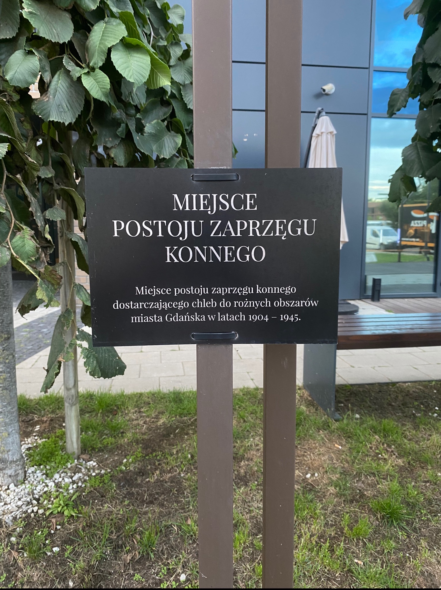 Tablica informacyjna: Miejsce postoju zaprzęgu konnego dostarczającego chleb w Gdańsku w latach 1904-1945. Tło: zieleń, budynek z monitoringiem.