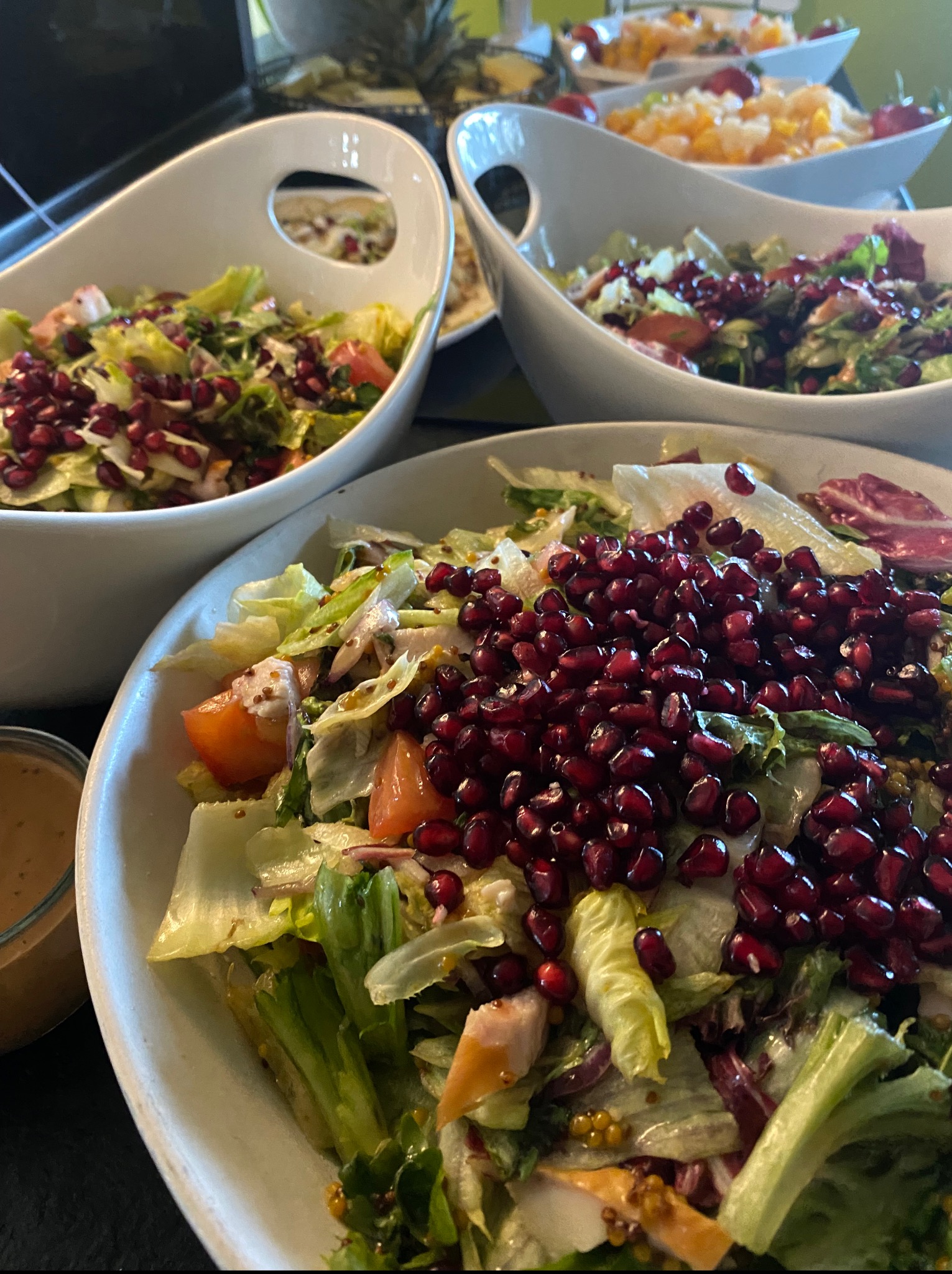 Elegancko podane sałatki z granatem w białych misach, ustawione na czarnym blacie. Widoczne inne potrawy w tle, sugerujące catering lub bufet.