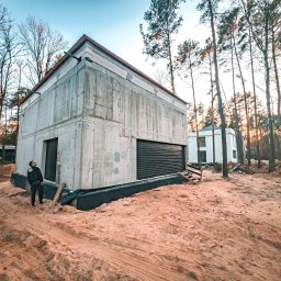 VITRU Construction - Surowy, betonowy dom w lesie, z widoczną konstrukcją i czarną bramą garażową. Mężczyzna w ciemnym ubraniu spogląda w górę na budynek. Piaszczyste podłoże.