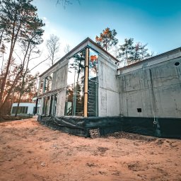VITRU Construction - Surowy, nowoczesny dom z betonu i szkła w lesie, w trakcie budowy. Widoczne szalunki i zabezpieczenia fundamentów. Nierówny teren, drzewa odbijają się w szybach.