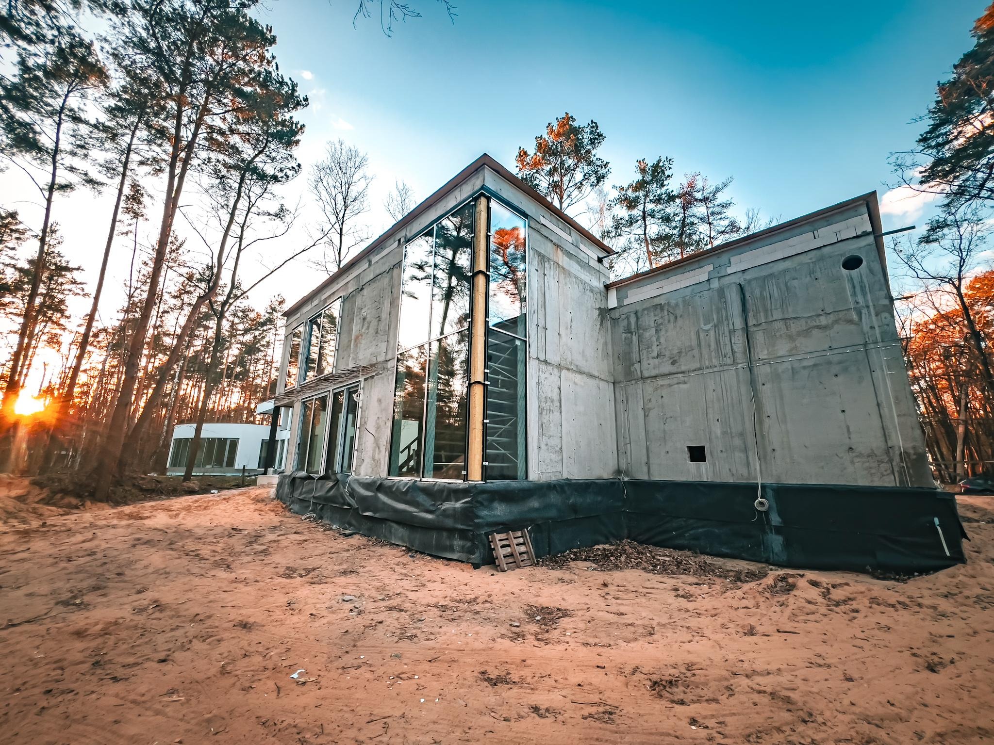 Surowy, nowoczesny dom z betonu i szkła w lesie, w trakcie budowy. Widoczne szalunki i zabezpieczenia fundamentów. Nierówny teren, drzewa odbijają się w szybach.