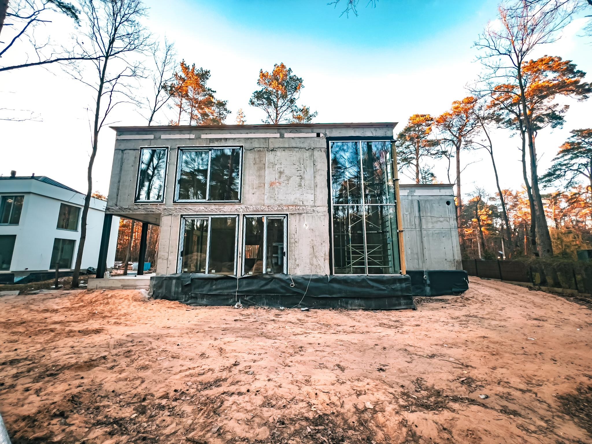 Surowy, betonowy dom w trakcie budowy, z widocznymi oknami i rusztowaniami, otoczony piaszczystym terenem i drzewami. Widok z dołu podkreśla surowość konstrukcji.