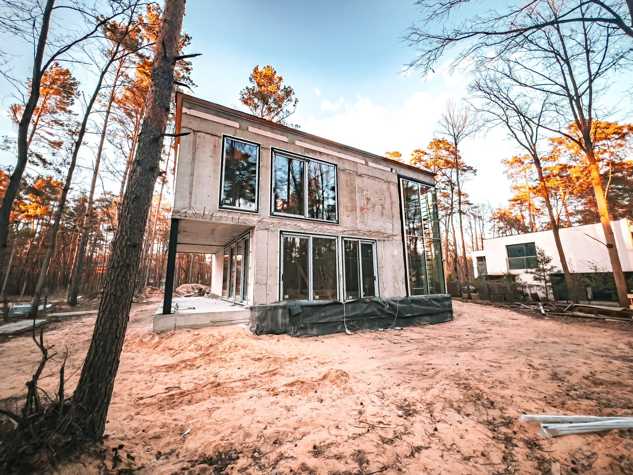 Surowy, dwukondygnacyjny dom z betonu architektonicznego w lesie, z dużymi oknami i widoczną konstrukcją. Etap budowy, piasek na działce, drzewa w tle.