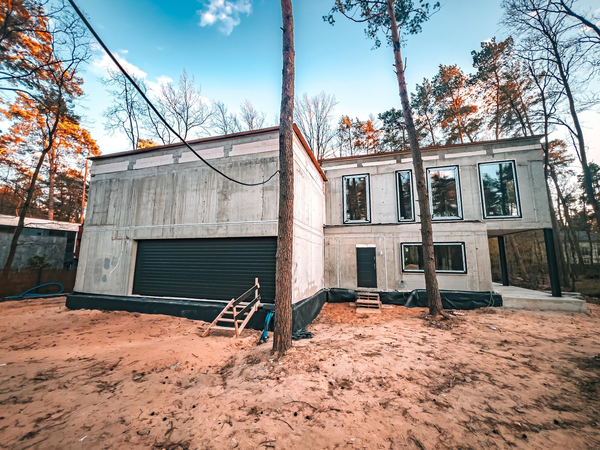 Surowy, dwukondygnacyjny dom z betonu komórkowego w lesie, z czarną bramą garażową i oknami w czarnych ramach. Widoczne drewniane schody i drzewa na pierwszym planie.