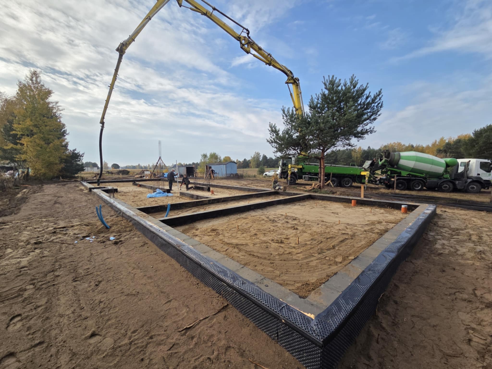 Wylane fundamenty domu jednorodzinnego z użyciem pompy do betonu. Widoczny mikser betonu i pracownicy budowlani. Fundamenty zabezpieczone folią kubełkową.
