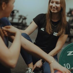 Natalia Czarnecka Trenerka Kobiet - Uśmiechnięta kobieta w czarnej koszulce z logo Just Gym, siedząca na podłodze siłowni, pomaga klientce w ćwiczeniach. W tle obciążenia i sztanga.