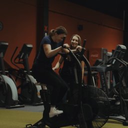 Natalia Czarnecka Trenerka Kobiet - Uśmiechnięta kobieta ćwiczy na rowerze powietrznym w siłowni, z drugą osobą obok. Sprzęt Matrix, pomarańczowa ściana w tle.