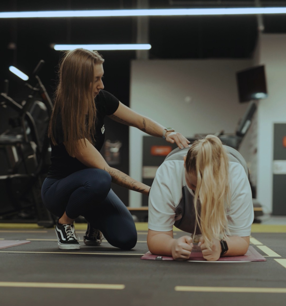 Kobieta w trakcie ćwiczeń z trenerką w siłowni. Trenerka koryguje postawę ćwiczącej. Widoczne maty do ćwiczeń i sprzęt fitness w tle.