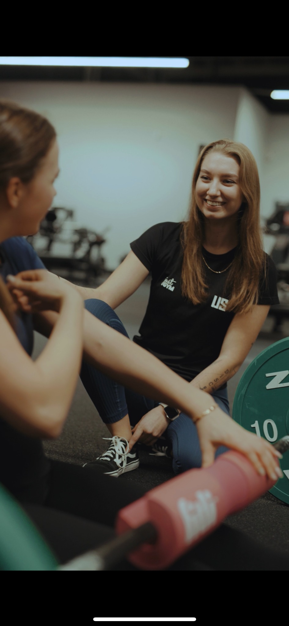 Uśmiechnięta kobieta w czarnej koszulce z logo Just Gym, siedząca na podłodze siłowni, pomaga klientce w ćwiczeniach. W tle obciążenia i sztanga.