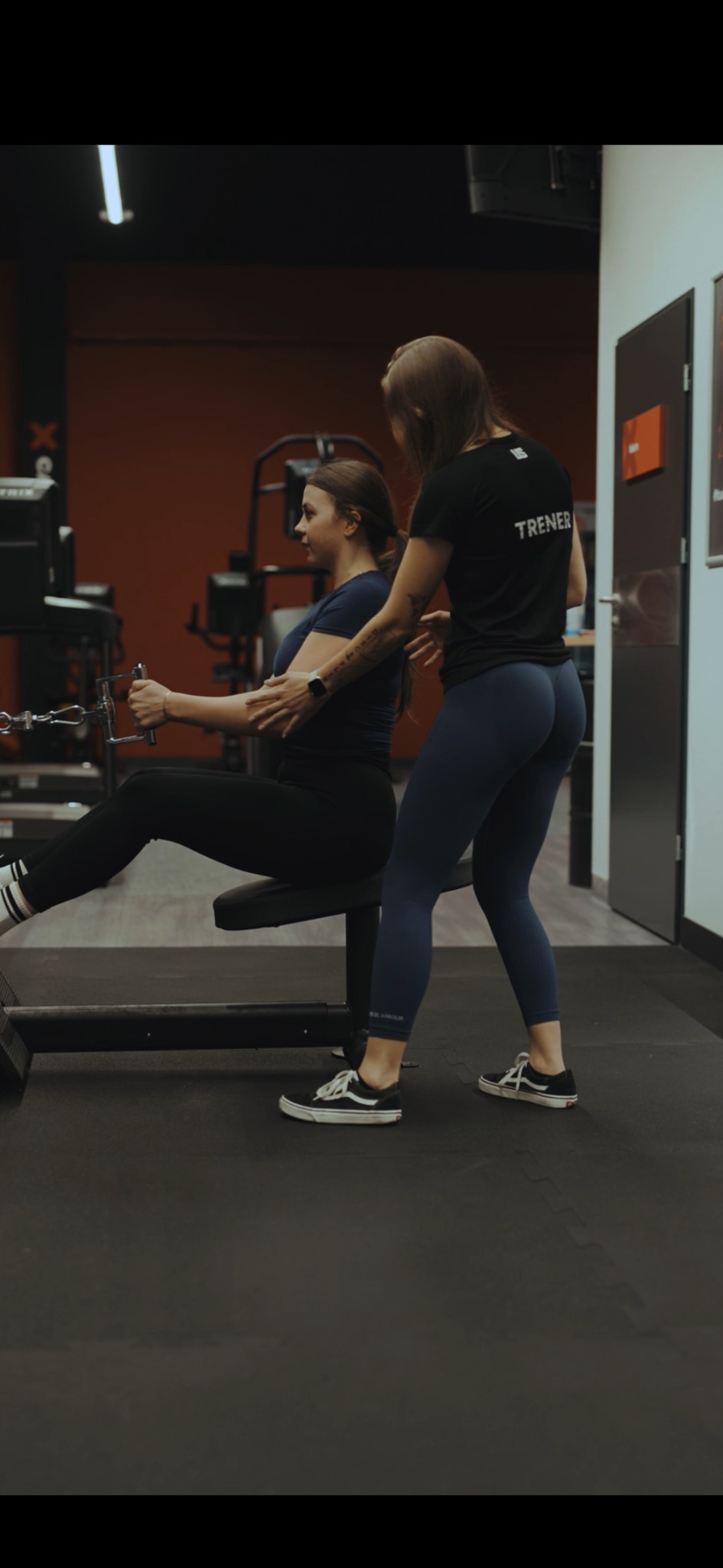 Kobieta ćwiczy na maszynie pod okiem trenerki w czarnej koszulce z napisem 'TRENER' w siłowni z czerwonym akcentem na ścianie. Ćwiczenie na mięśnie pleców.
