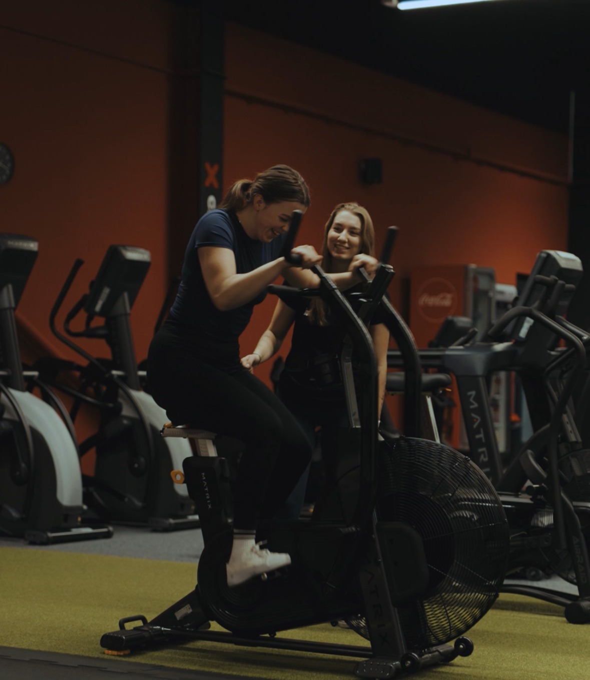 Uśmiechnięta kobieta ćwiczy na rowerze powietrznym w siłowni, z drugą osobą obok. Sprzęt Matrix, pomarańczowa ściana w tle.