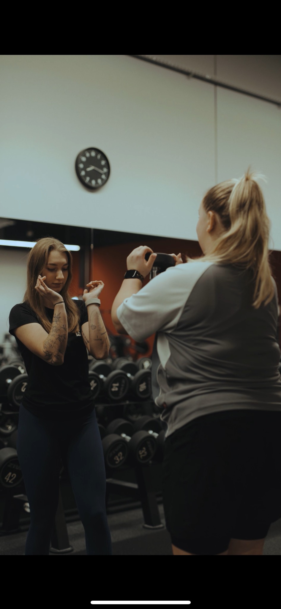 Kobieta z tatuażami ćwiczy z hantlami pod okiem drugiej kobiety w siłowni. W tle regał z hantlami i zegar.