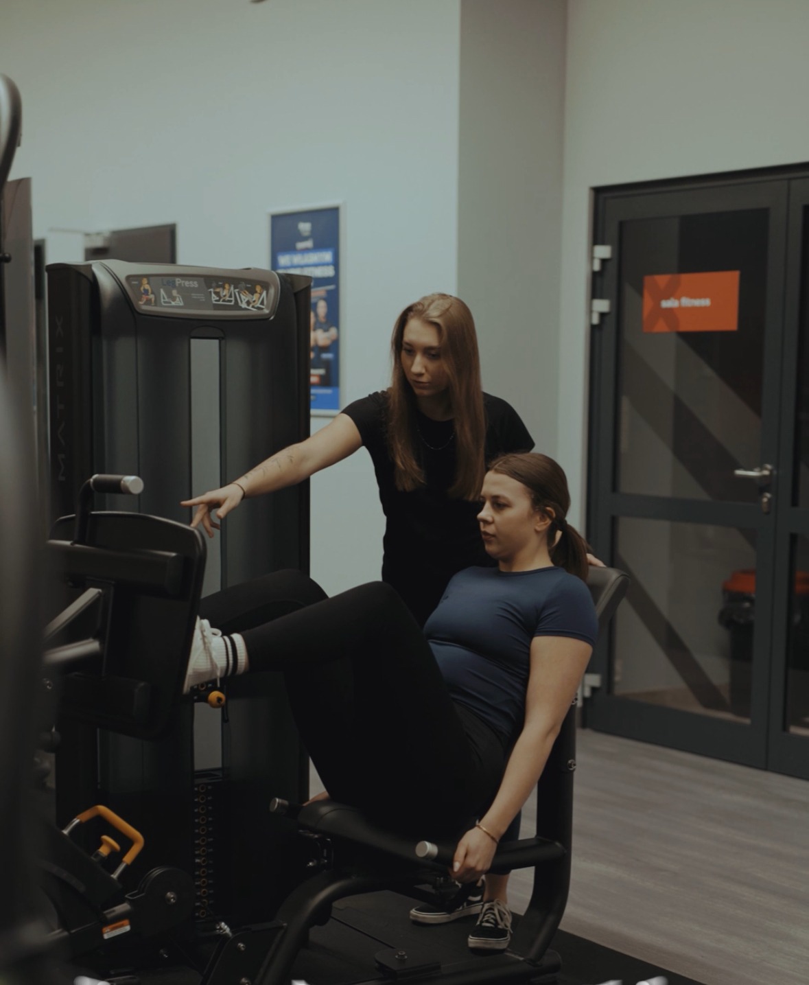 Kobieta ćwiczy na maszynie do wyciskania nóg pod okiem instruktorki w czarnej koszulce. Sala fitness z szarymi ścianami i ciemnymi drzwiami.