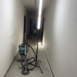 Perfekt Finish Sebastian Zdeb - Szlifierka do gładzi, odkurzacz przemysłowy i lampa budowlana w korytarzu podczas prac wykończeniowych. Widoczne ślady szlifowania na ścianach i podłodze.