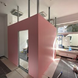 Perfekt Finish Sebastian Zdeb - Różowa ścianka działowa z otworem drzwiowym w trakcie budowy, widoczne profile metalowe i narzędzia. W tle okno z widokiem na ulicę.