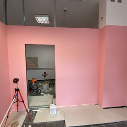 Perfekt Finish Sebastian Zdeb - Różowe ściany w trakcie prac wykończeniowych. Widoczny laser krzyżowy, narzędzia i materiały budowlane. W tle otwór w ścianie z widocznymi cegłami.