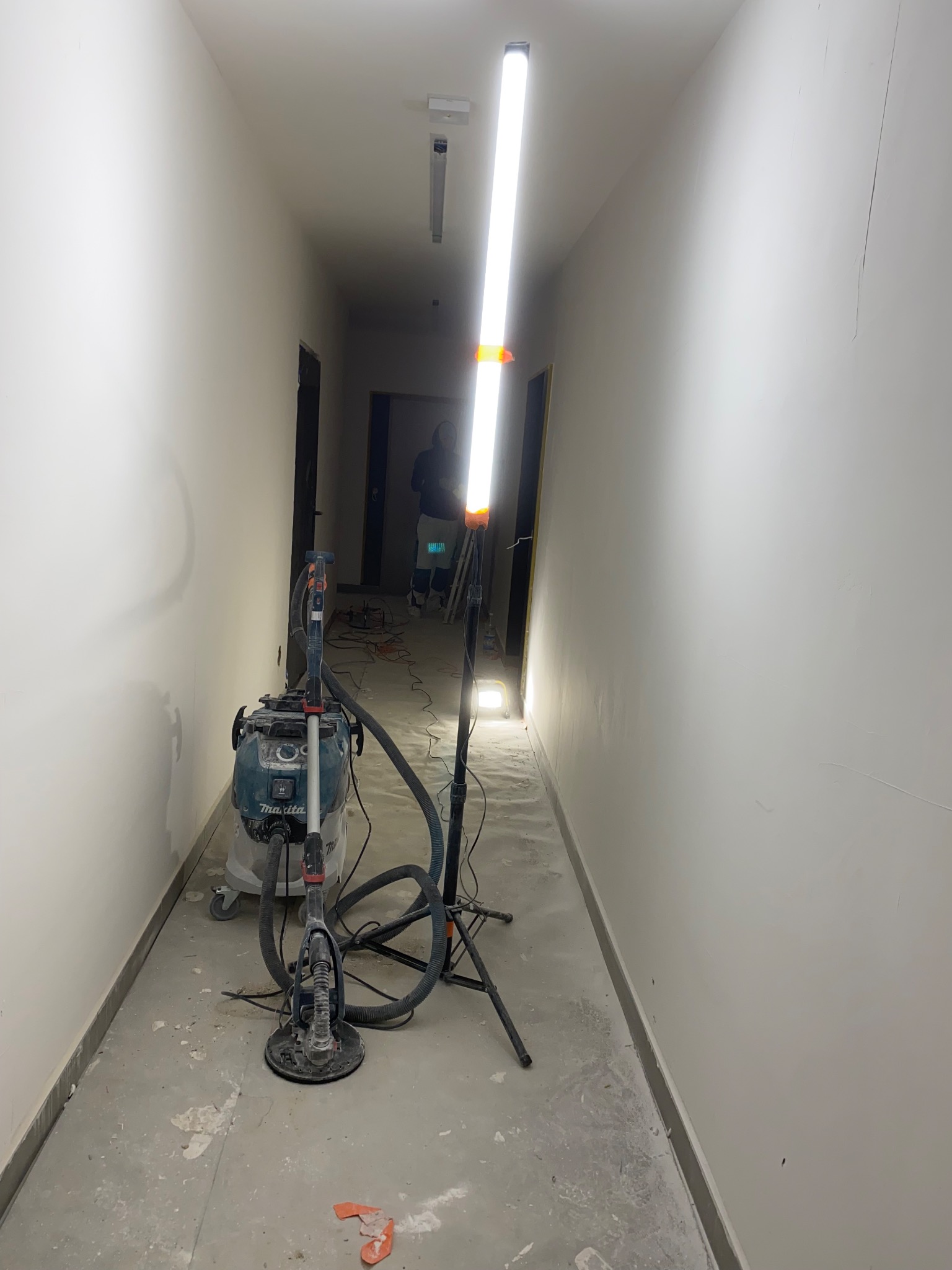 Szlifierka do gładzi, odkurzacz przemysłowy i lampa budowlana w korytarzu podczas prac wykończeniowych. Widoczne ślady szlifowania na ścianach i podłodze.