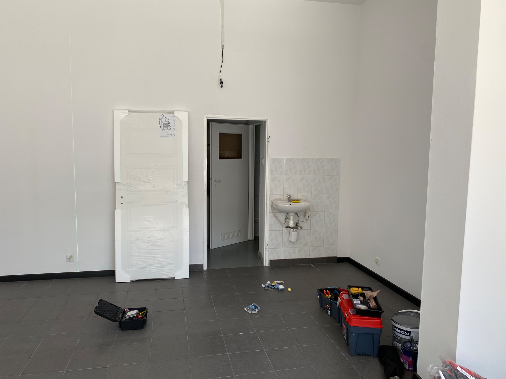 Wnętrze w trakcie remontu: białe ściany, szara podłoga, drzwi w folii, narzędzia i farby. Widoczna umywalka i laser krzyżowy. Przygotowania do wykończenia.