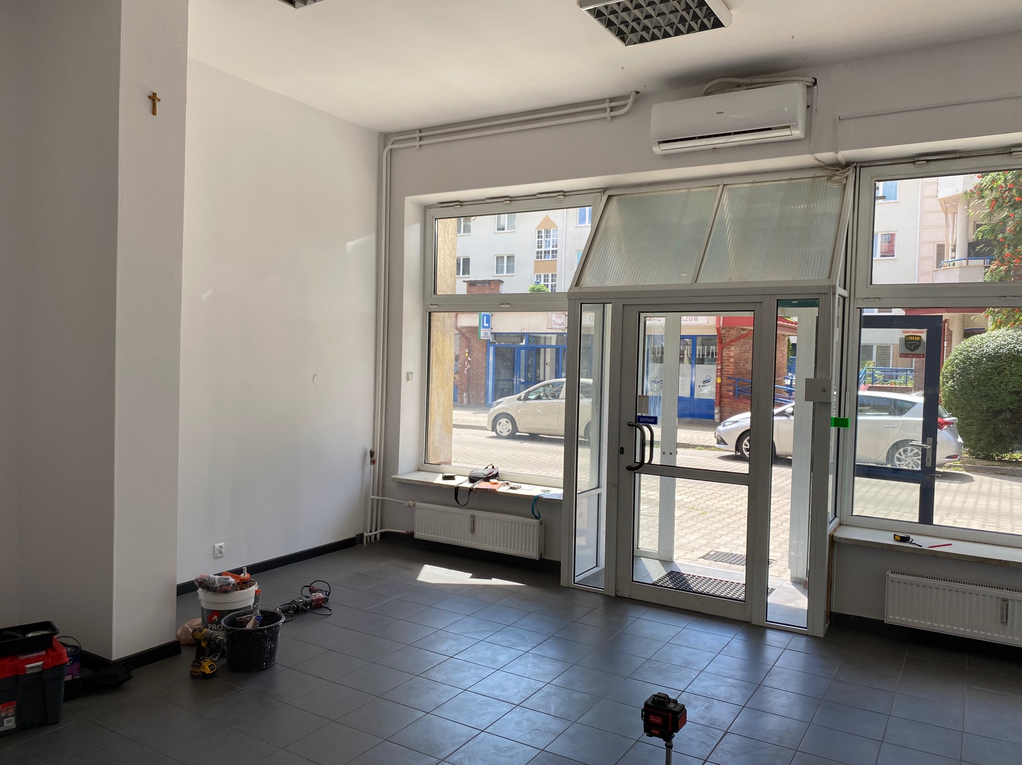 Jasne wnętrze lokalu przed pracami wykończeniowymi: szare płytki, białe ściany, okna z widokiem na ulicę. Widoczne narzędzia i materiały malarskie.