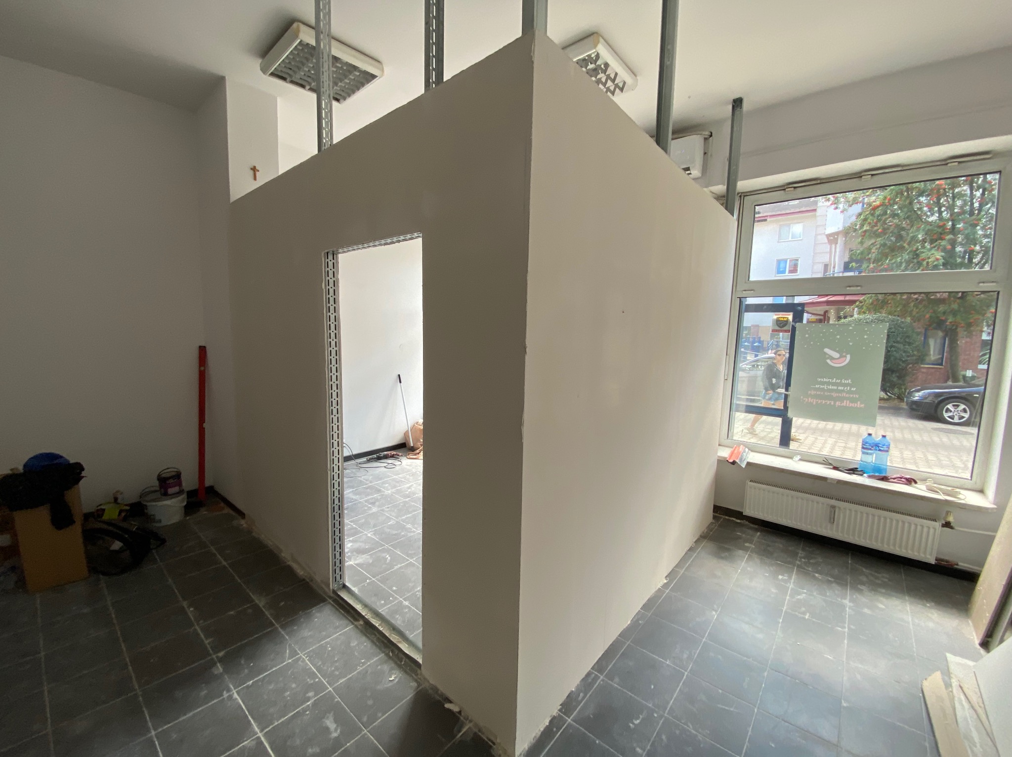 Świeżo pomalowana ściana działowa z otworem drzwiowym w jasnym pomieszczeniu z szarą podłogą. Widoczne metalowe profile sufitowe i narzędzia malarskie.