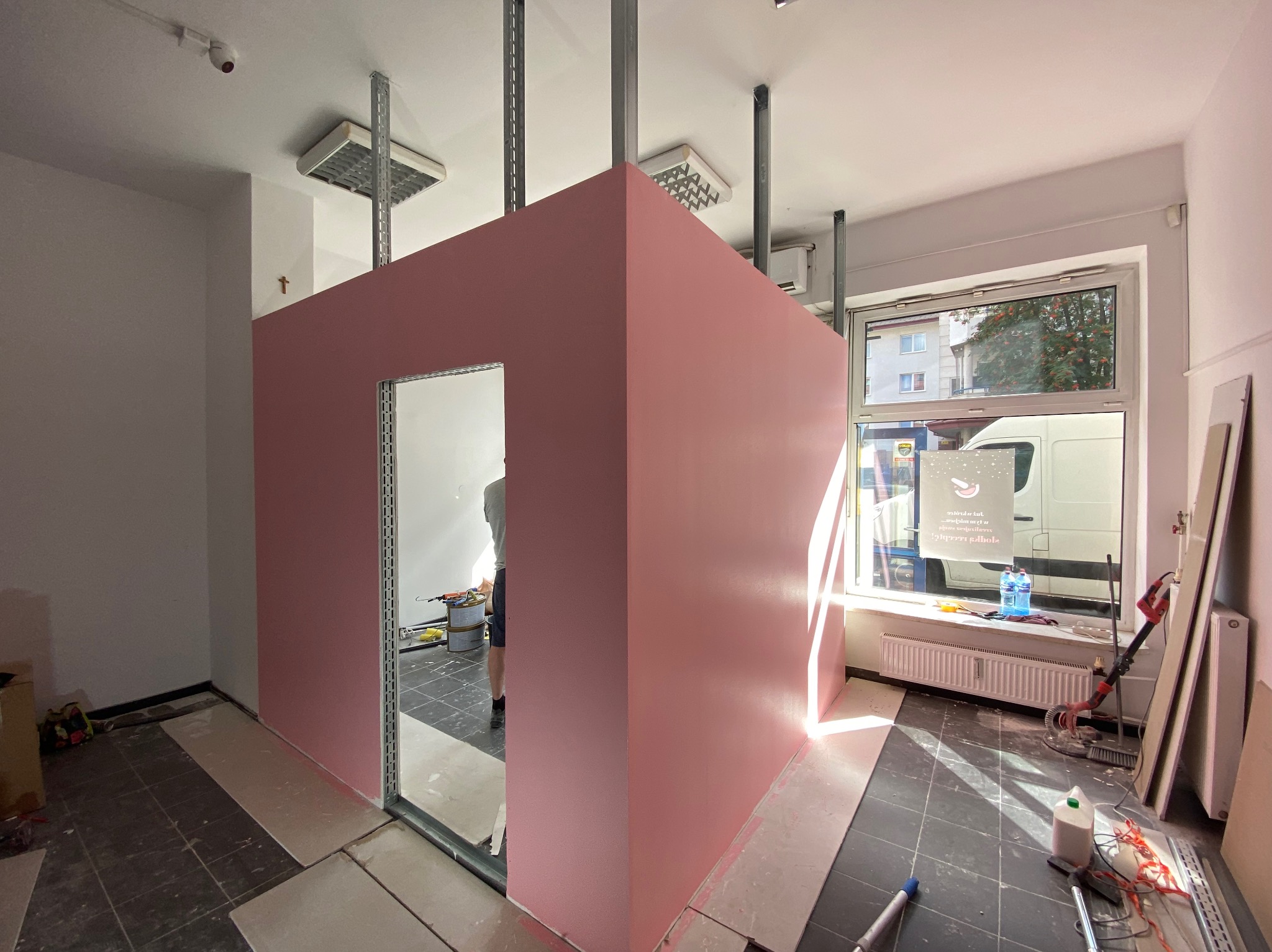 Różowa ścianka działowa z otworem drzwiowym w trakcie budowy, widoczne profile metalowe i narzędzia. W tle okno z widokiem na ulicę.