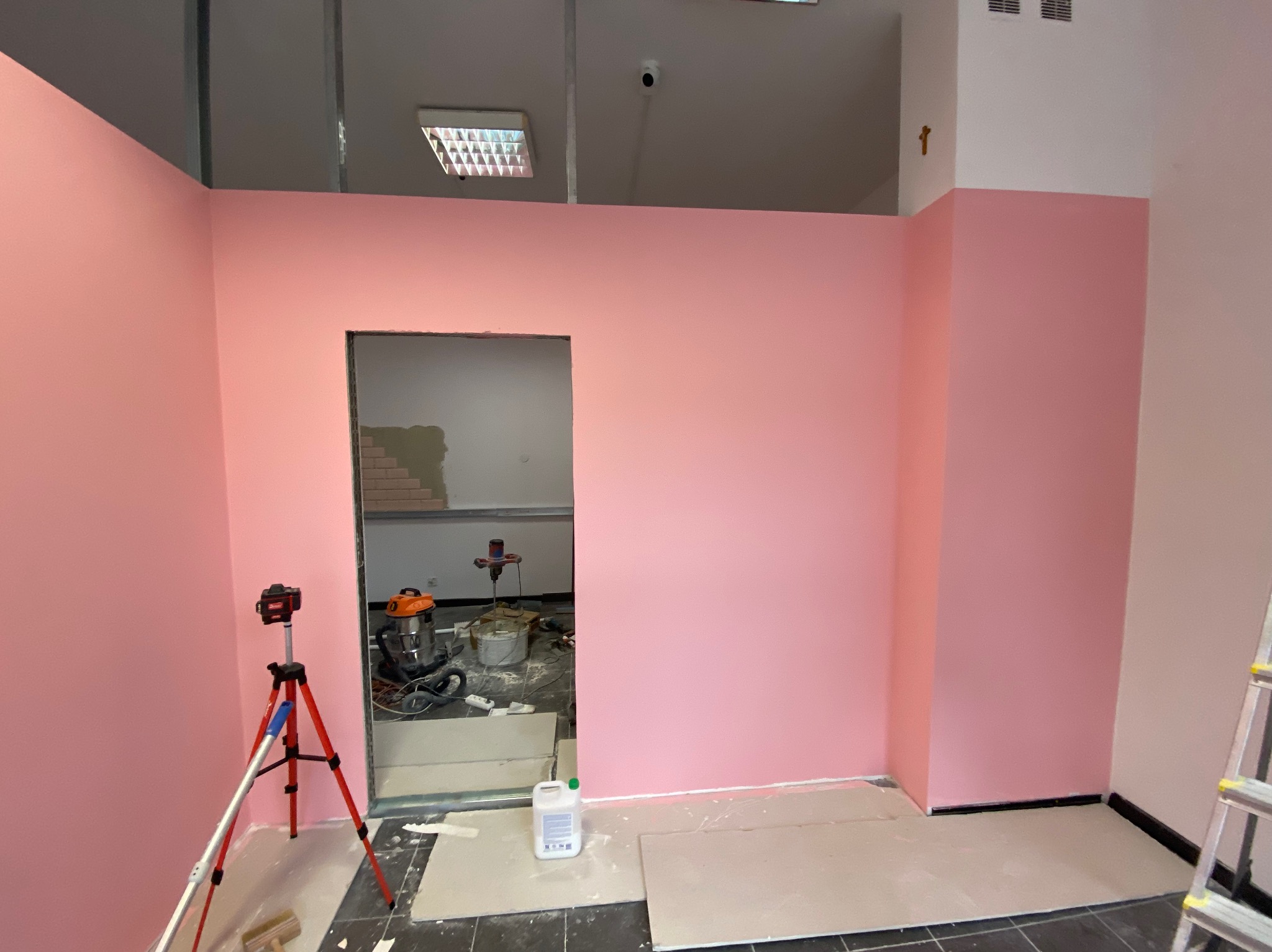 Różowe ściany w trakcie prac wykończeniowych. Widoczny laser krzyżowy, narzędzia i materiały budowlane. W tle otwór w ścianie z widocznymi cegłami.