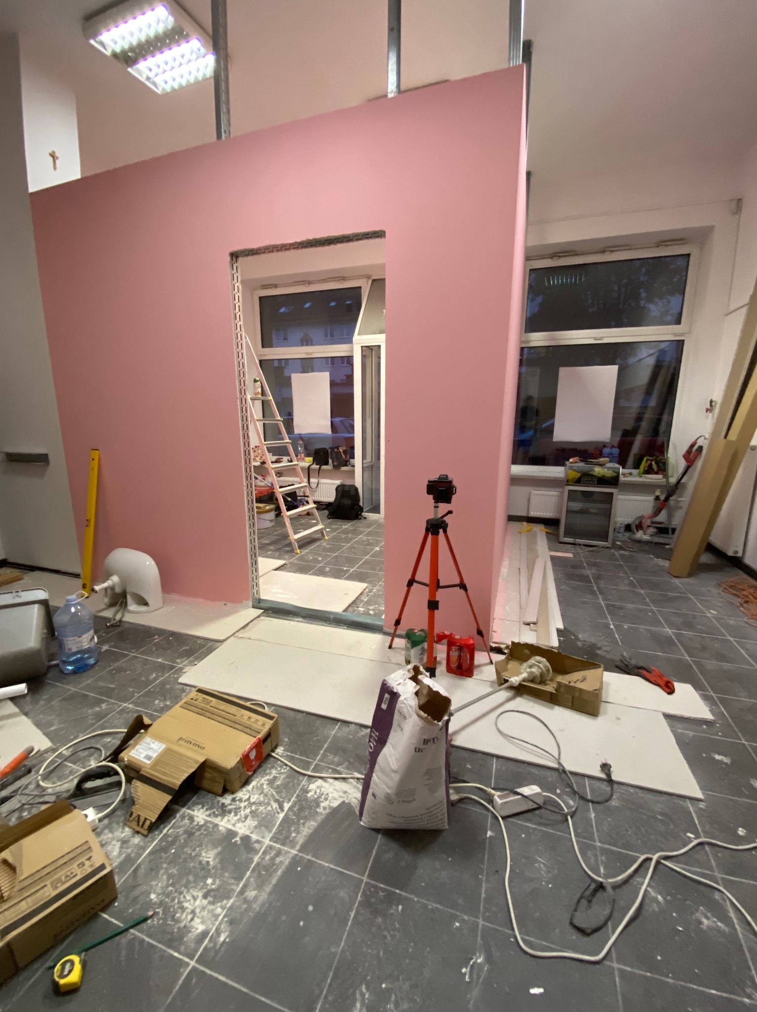 Różowa ściana z otworem drzwiowym w trakcie budowy, widoczne profile metalowe, narzędzia i materiały budowlane na podłodze. W tle okno i drabina.