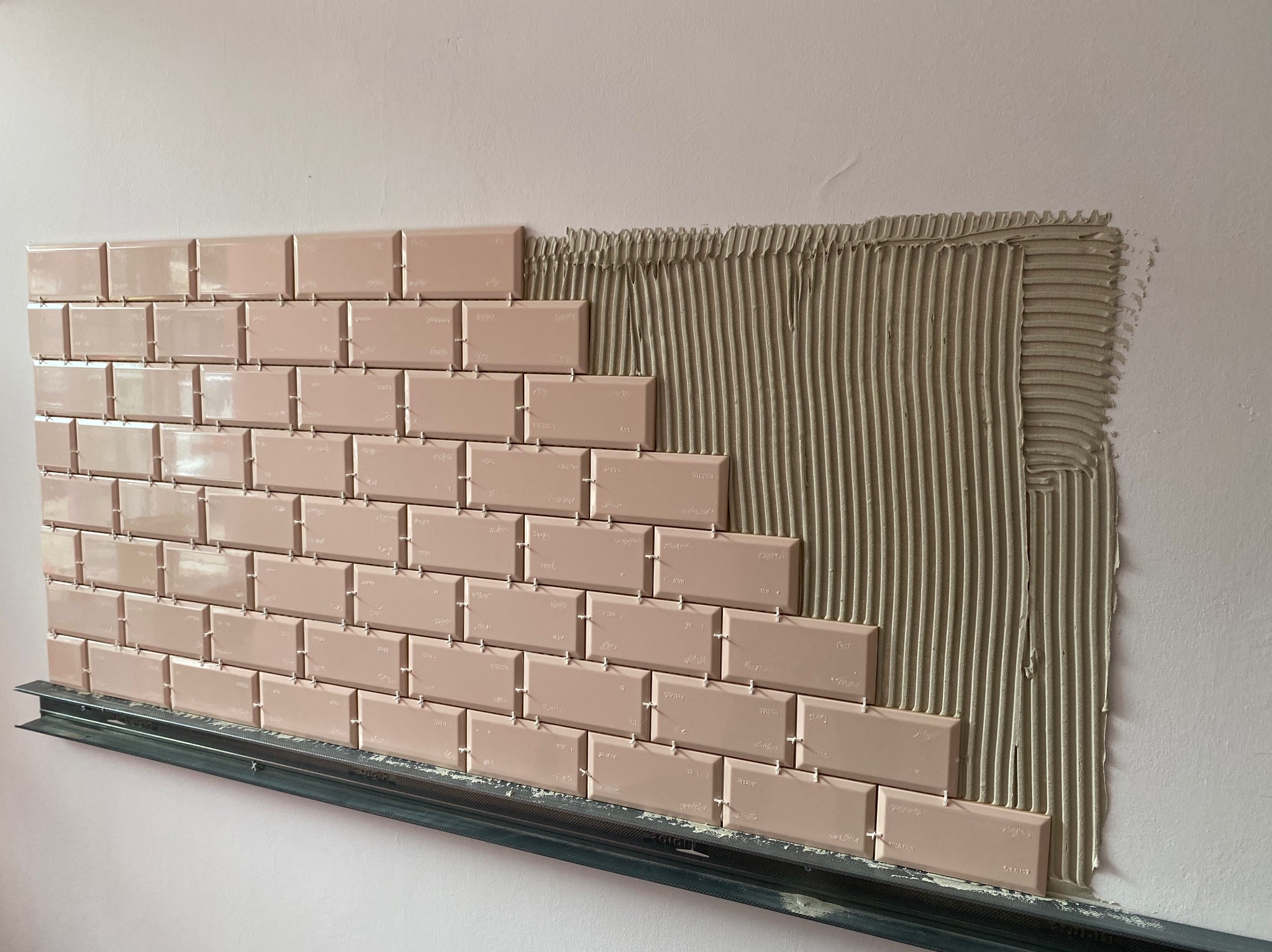 Układanie beżowych, prostokątnych płytek na ścianie. Widoczny fragment ściany z nałożonym klejem i metalowa listwa podtrzymująca płytki. Etap prac glazurniczych.