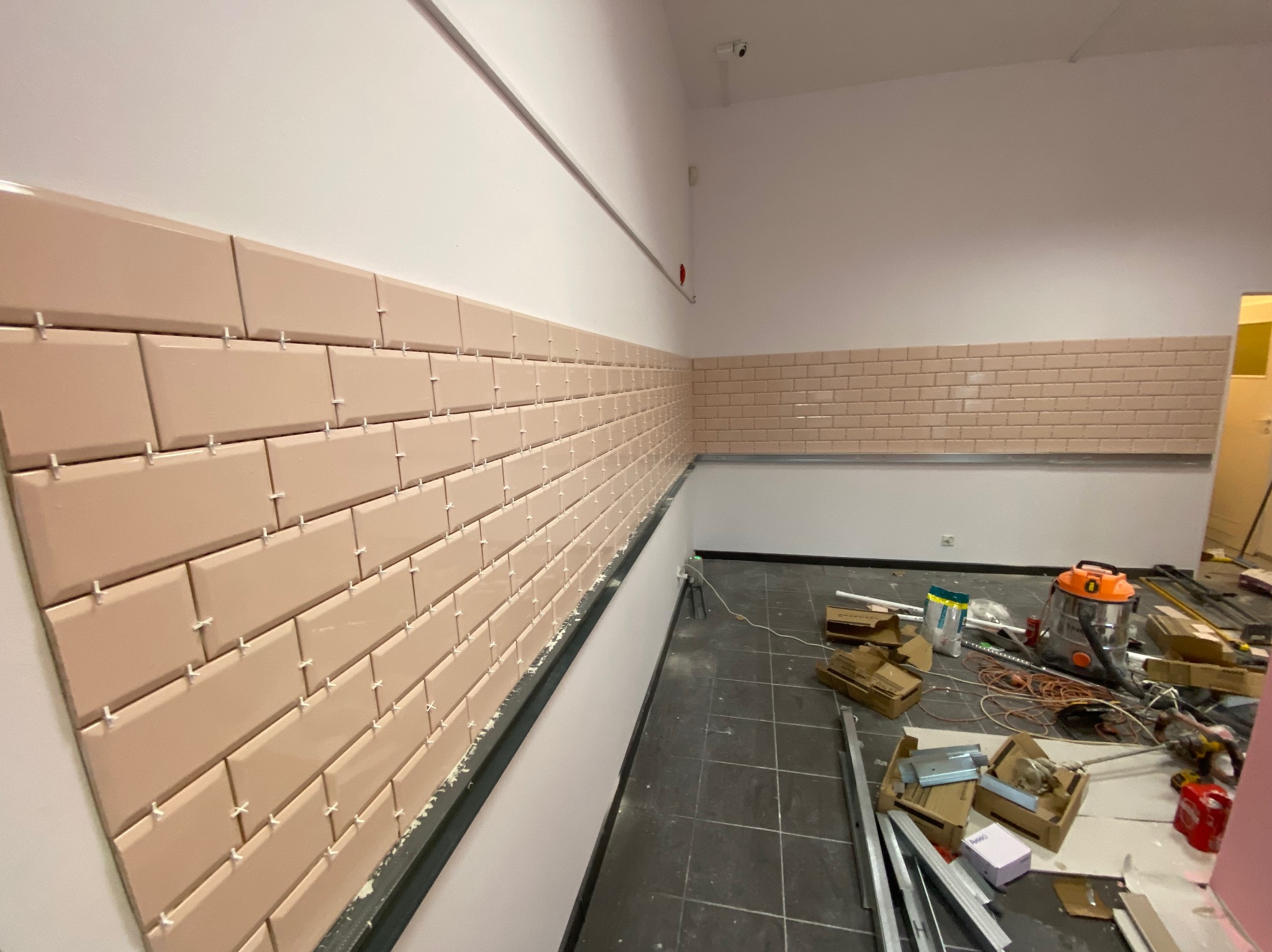 Układanie beżowych, prostokątnych płytek na ścianie z użyciem krzyżyków dystansowych. Widoczne narzędzia i materiały budowlane na podłodze. Prace glazurnicze w toku.