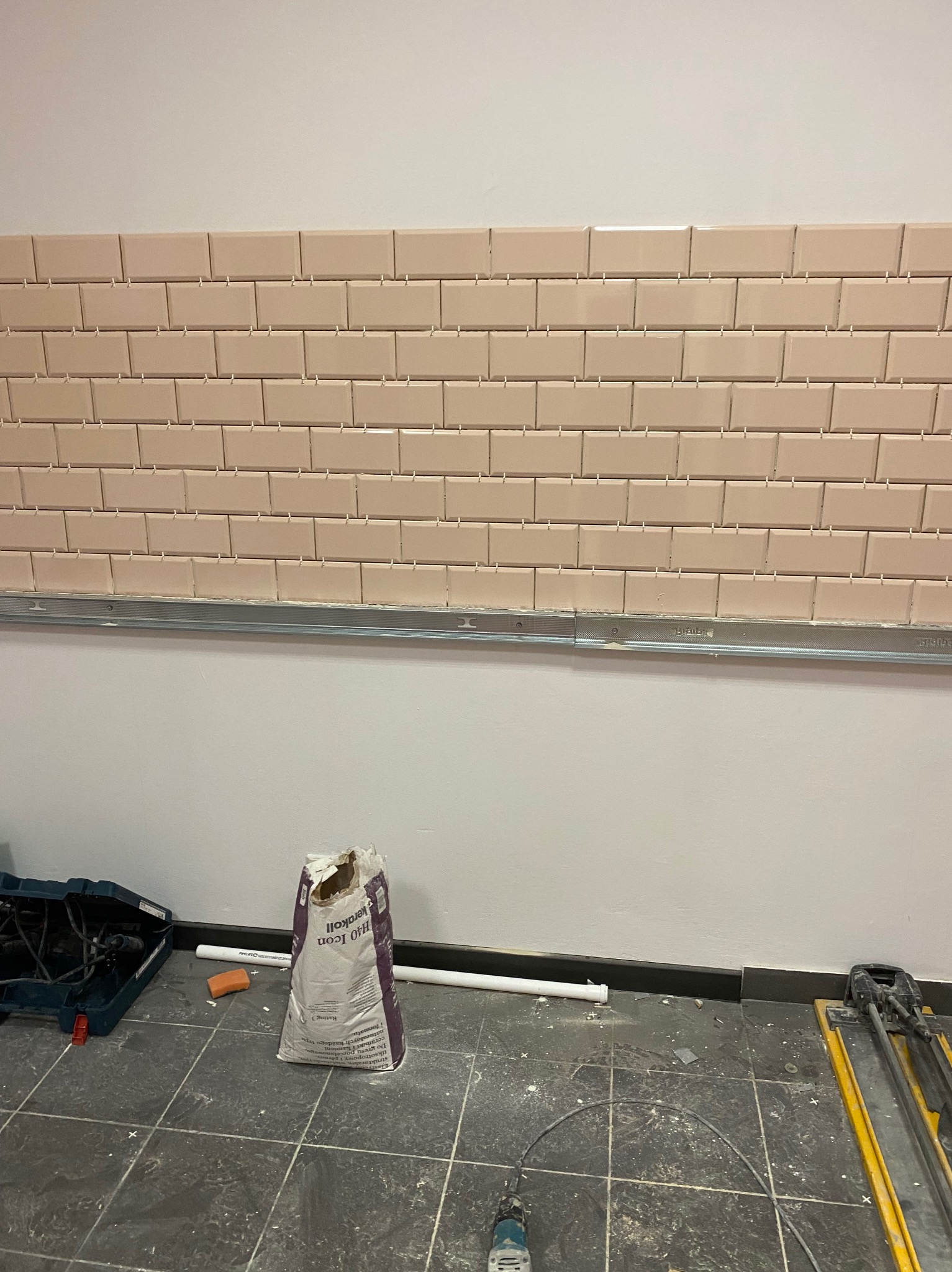 Ściana z beżowych, prostokątnych płytek, częściowo ułożonych, z widocznymi narzędziami i materiałami budowlanymi na podłodze. Etap prac wykończeniowych.