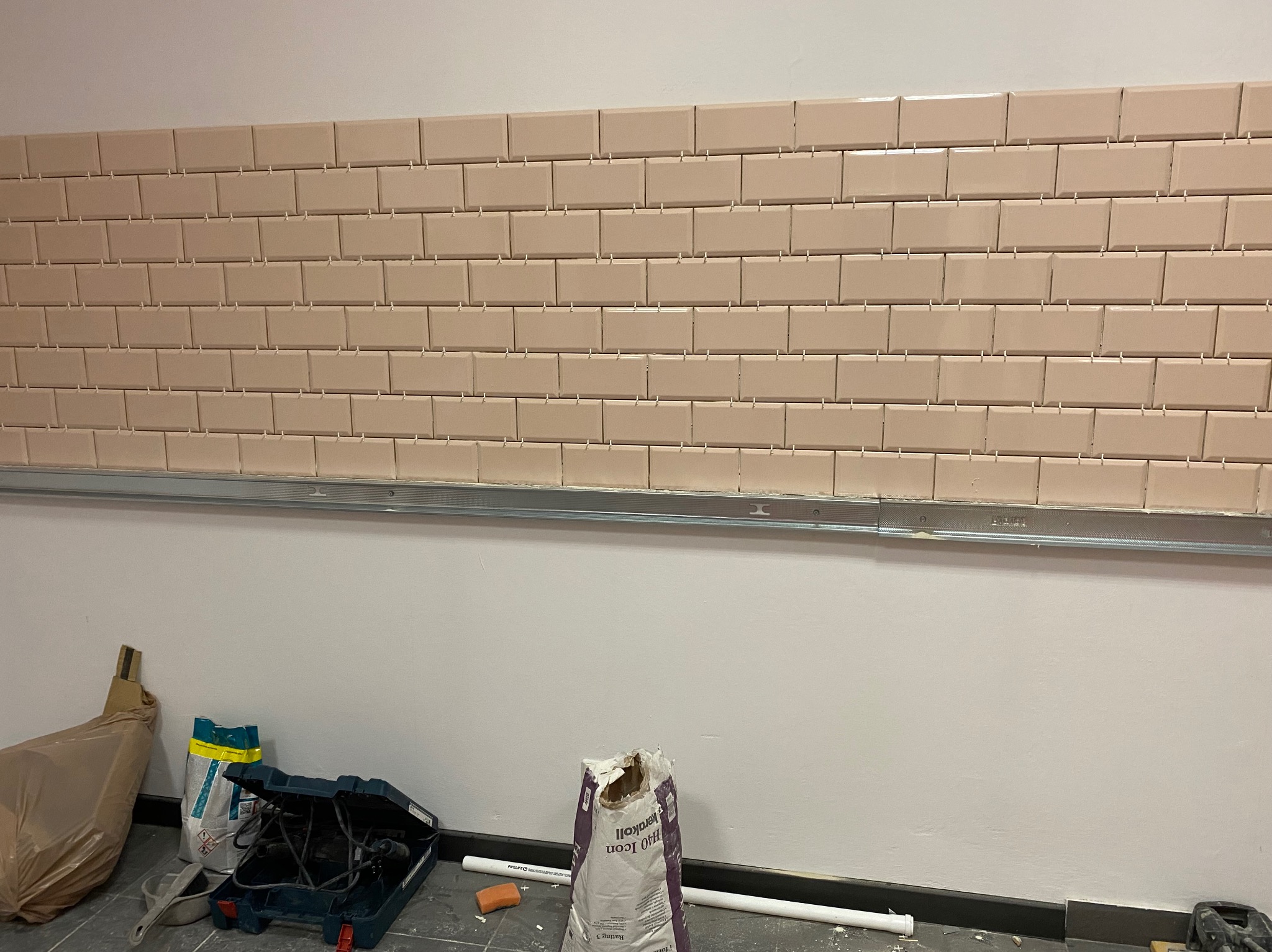 Ściana z beżowych, prostokątnych płytek, ułożonych w cegiełkę, z widocznymi krzyżykami dystansowymi. Na podłodze narzędzia i worki z zaprawą.