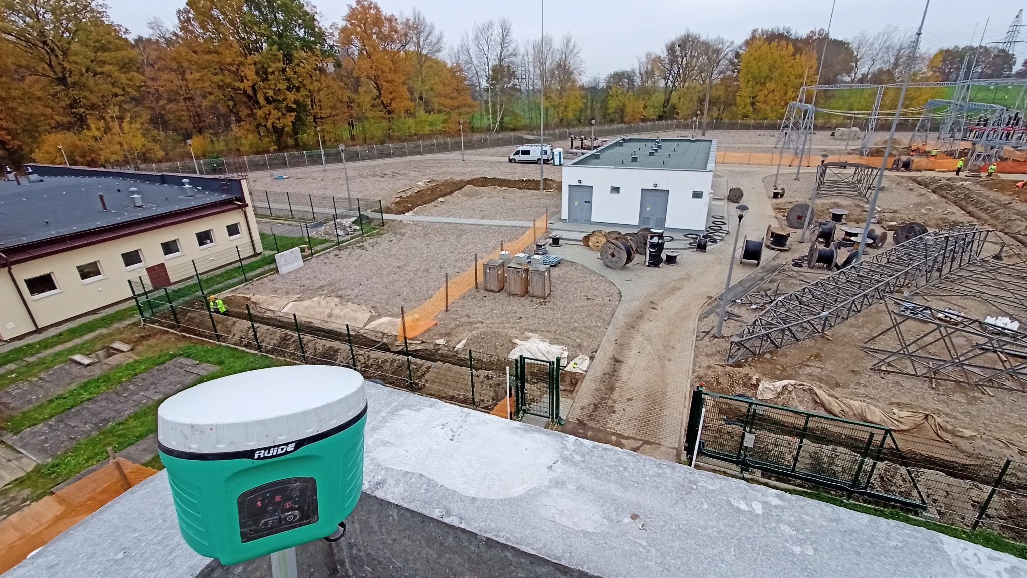 Prace geodezyjne na terenie budowy: widok z góry na zielony odbiornik GNSS Ruide, teren z wykopami, budynki i konstrukcje stalowe w tle. Jesienna sceneria.