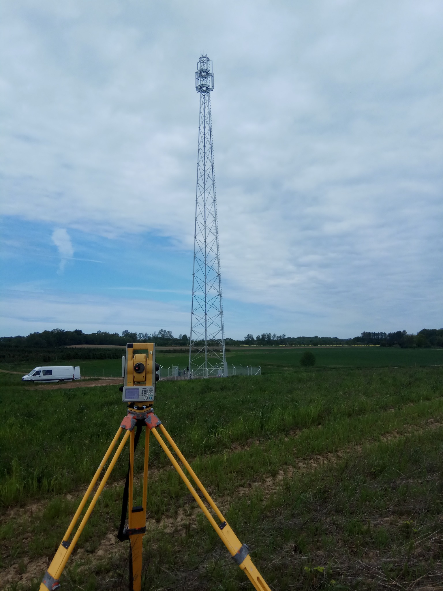 Żółty teodolit na trójnogu na tle zielonego pola i wieży telekomunikacyjnej pod błękitnym niebem z chmurami. W tle biały bus.
