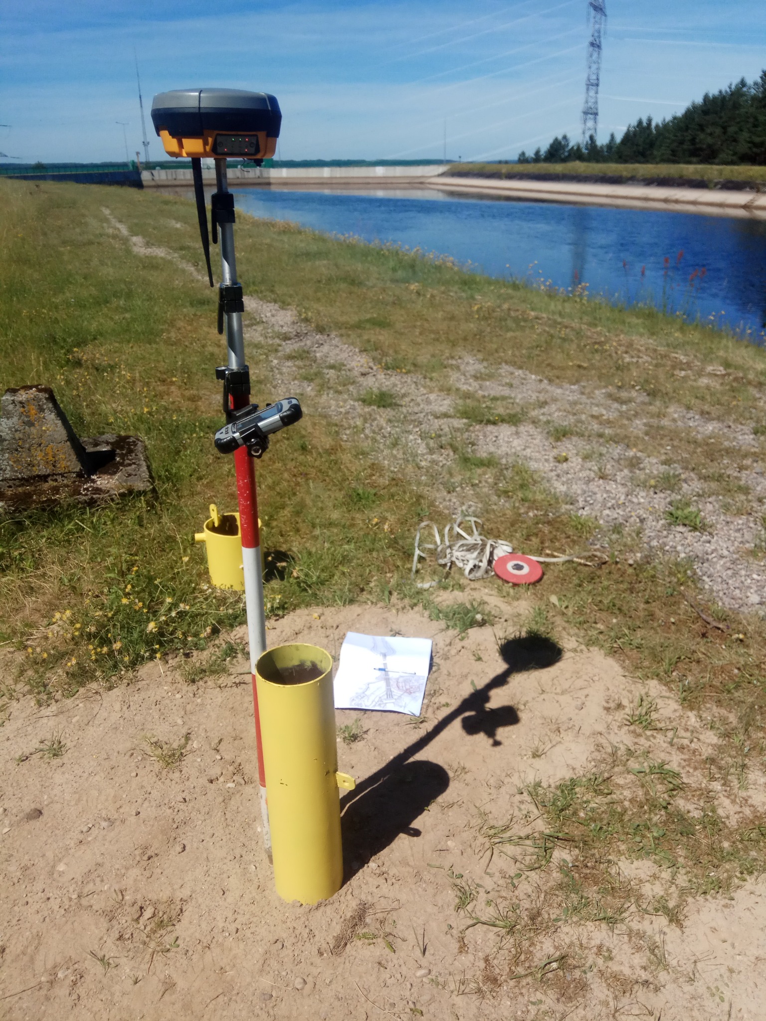Praca geodezyjna w terenie: tyczka geodezyjna z odbiornikiem GPS, żółta osłona, mapa i akcesoria na tle kanału wodnego i linii wysokiego napięcia.