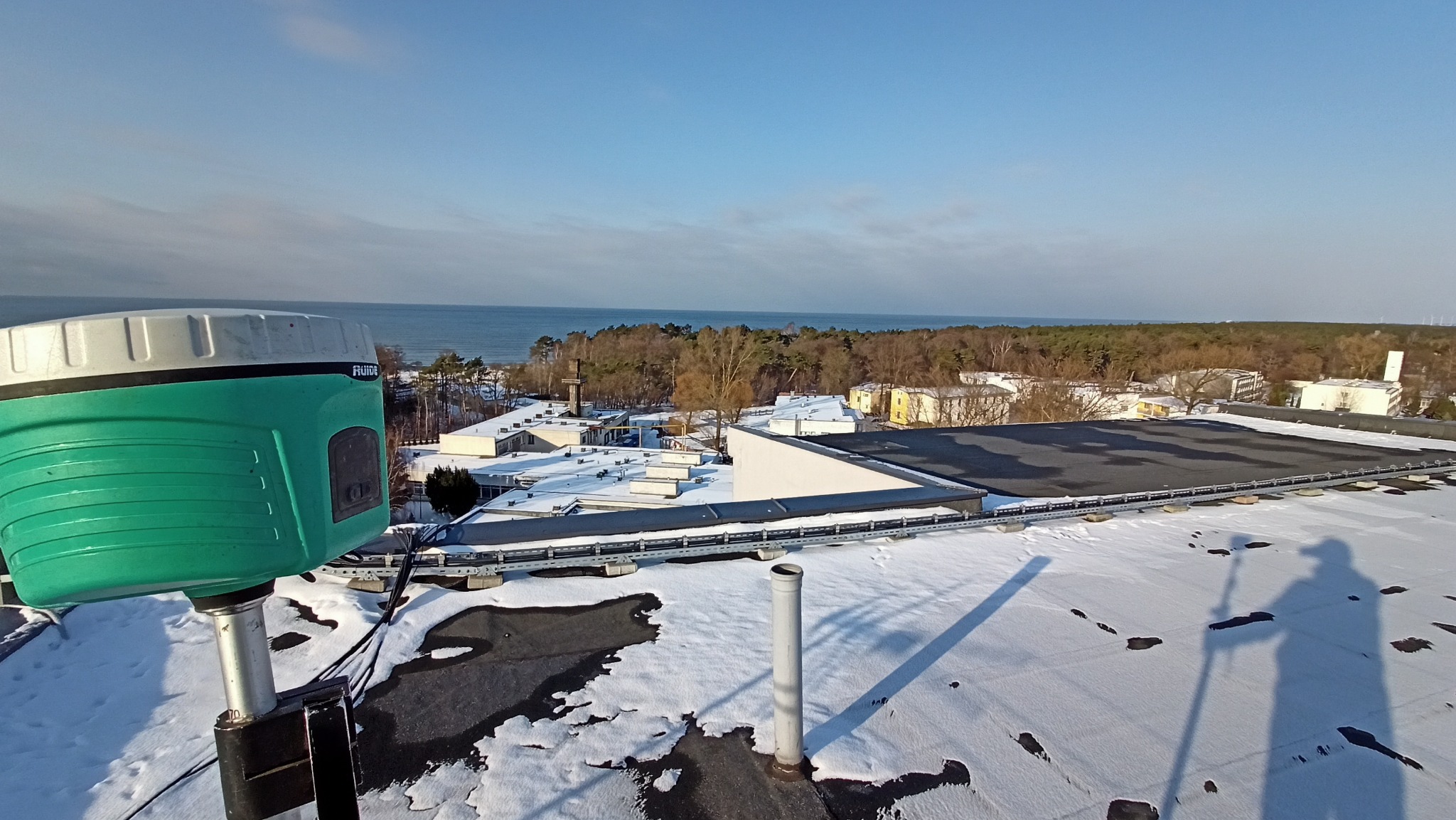 Geodezyjny odbiornik GNSS marki Ruide na dachu budynku pokrytego śniegiem, w tle widok na morze i las. Cień osoby obsługującej urządzenie.
