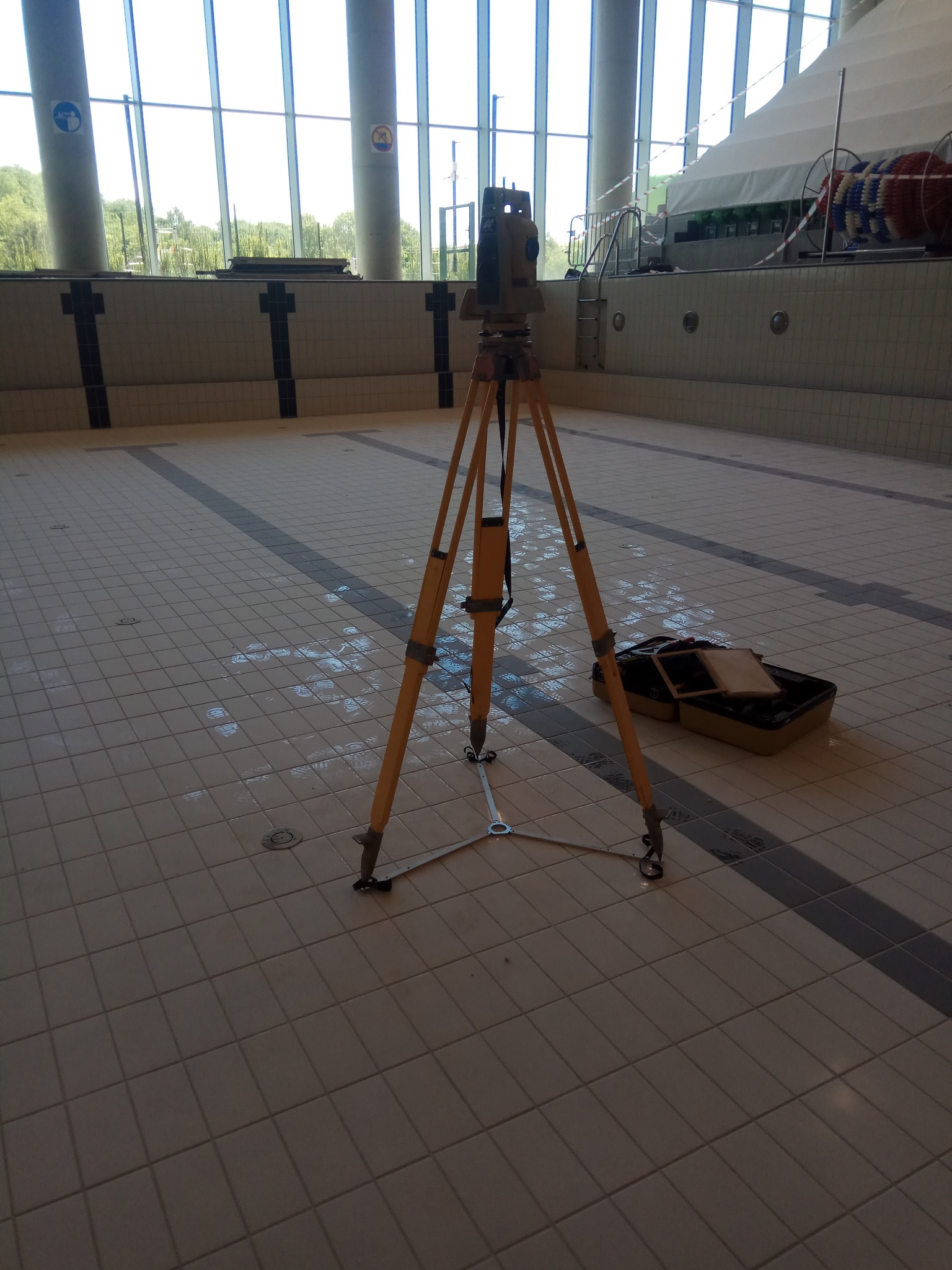 Tachimetr na trójnogu w pustym basenie z białymi płytkami i dużymi oknami. Sprzęt geodezyjny ustawiony do pomiarów wewnątrz obiektu sportowego.