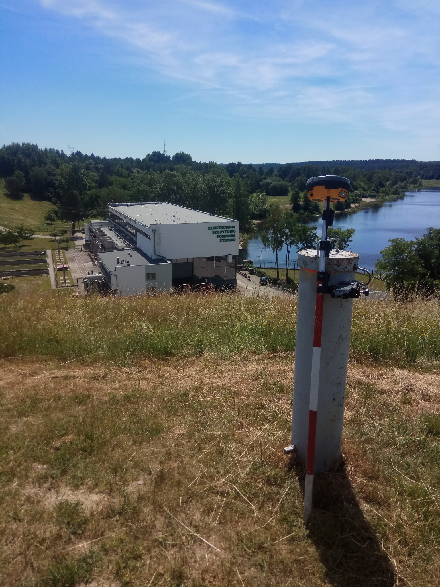 Pomiar geodezyjny z użyciem odbiornika GNSS na tle budynku elektrowni i jeziora, w słoneczny dzień. Widok z góry, na pierwszym planie tyczka geodezyjna.
