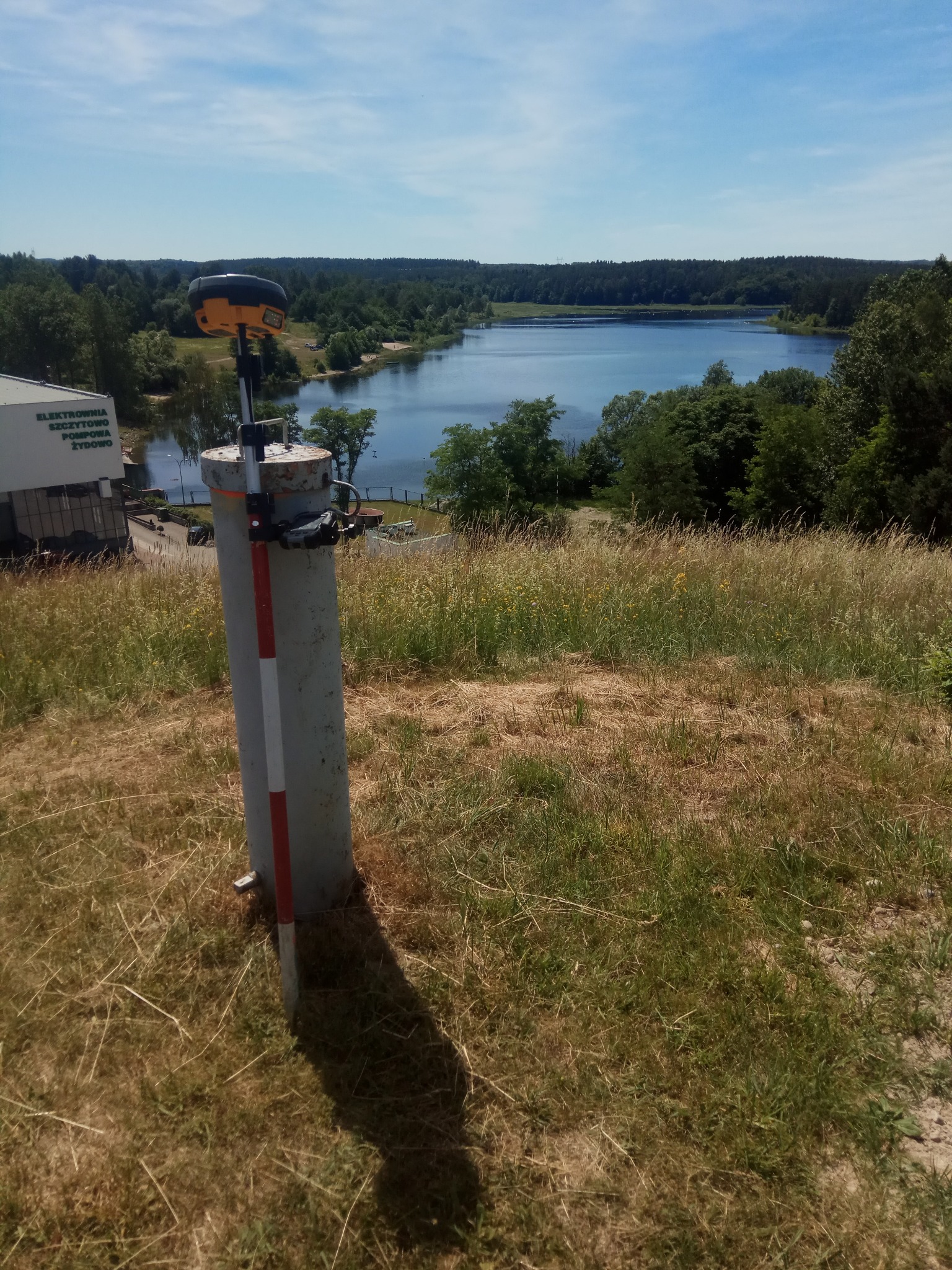 Przenośny odbiornik GNSS na tyczce geodezyjnej, umieszczony na betonowym słupku. W tle zbiornik wodny i zalesione tereny. Słoneczny dzień.