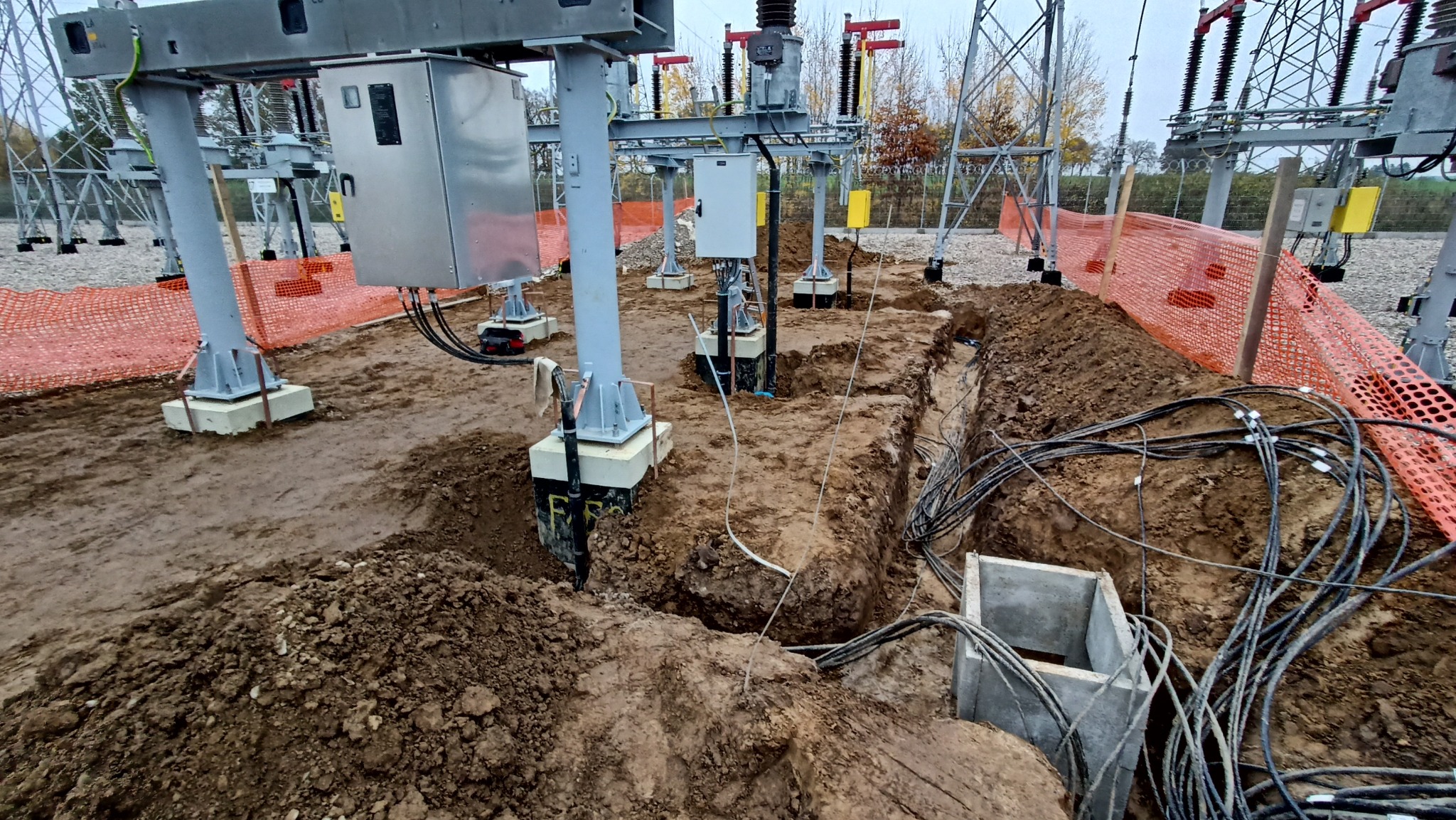 Budowa stacji transformatorowej: fundamenty pod słupy, wykopane rowy kablowe, pomarańczowa siatka zabezpieczająca teren. Prace ziemne w toku, widoczne kable.
