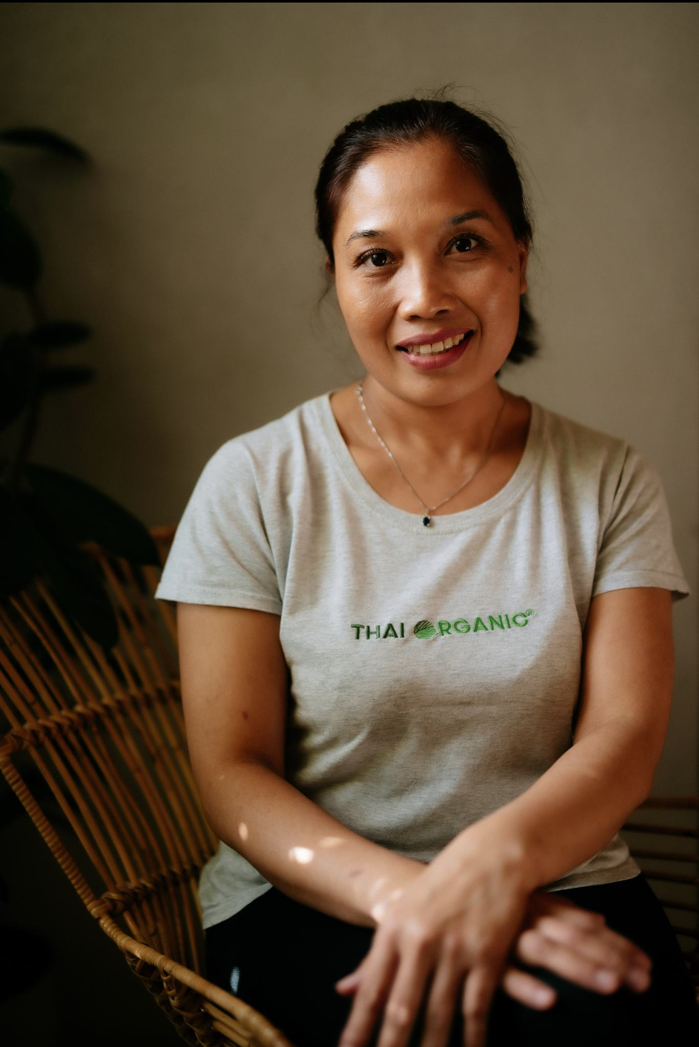 Uśmiechnięta kobieta w szarej koszulce z napisem 'Thai Organic' siedzi na wiklinowym krześle, w tle widoczna roślina doniczkowa i ściana w neutralnym odcieniu.