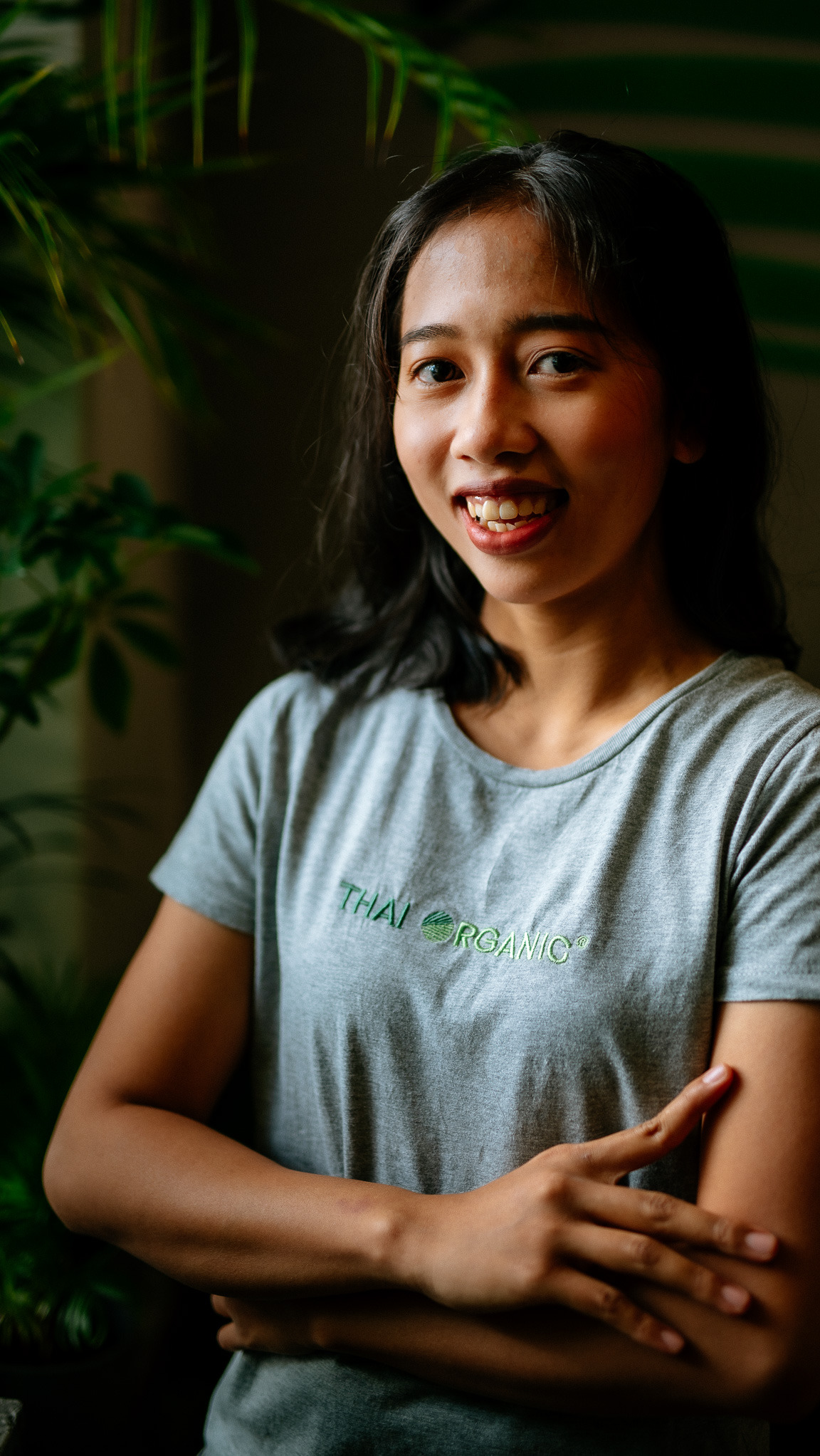 Uśmiechnięta kobieta z ciemnymi włosami w szarej koszulce z napisem 'Thai Organic', skrzyżowane ręce, zielone rośliny w tle, naturalne światło.