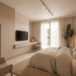 REDLICKI STUDIO Piotr Redlisz-Redlicki - Jasna sypialnia w beżach: duże łóżko, szafa w zabudowie, biurko z krzesłem, TV na ścianie, roślina doniczkowa i dywan z juty. Minimalistyczny design z naturalnym światłem.